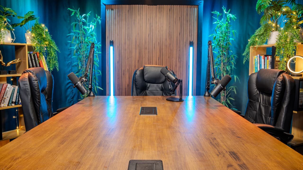 Boardroom at Bangkok Podcast Studio with a long wood table, black leather chairs, four microphone arms and blue lit walls.