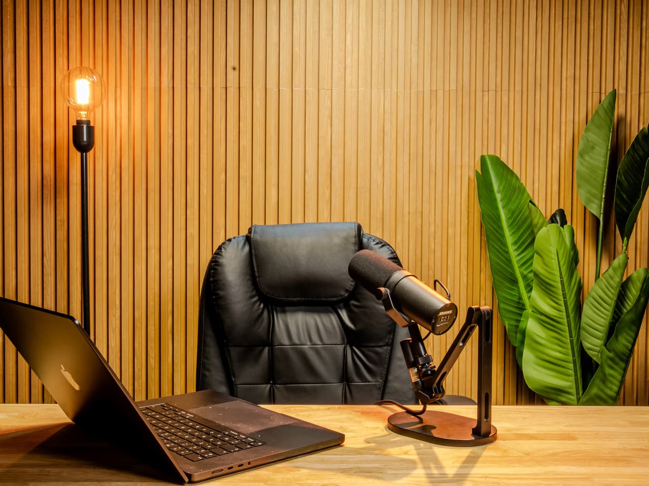 Desk at Bangkok Podcast Studio against a wood slat wall, with an open laptop, a microphone on a boom arm and a plant.