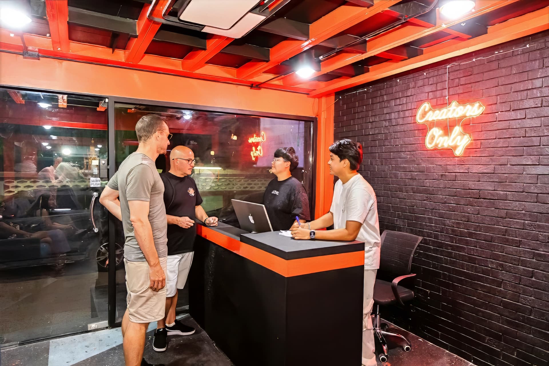 Reception desk at Bangkok Podcast Studio, black brick wall with an orange neon Creators Only sign, four people standing around a laptop on the counter.